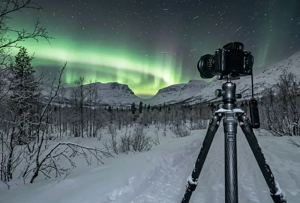 Abisko astrophotography gear: a mirrorless camera and wide-angle lens on a carbon-fiber tripod facing a green aurora curtain over a snow-covered birch forest, with faint star trails above the Lapporten mountains.