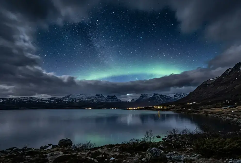 Wide-angle night photograph over Abisko National Park and Lake Torneträsk in Swedish Lapland: a sharply defined gap of clear, star-filled sky ringed by cloud at the horizon, illustrating Abisko's clear-sky microclimate, explained by the rain shadow cast by the Scandinavian Mountains, with a faint green aurora glow above the water.