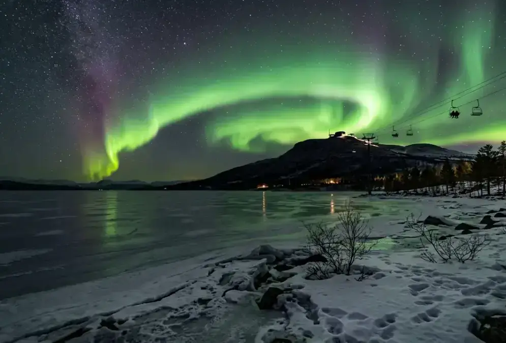 Aurora borealis over Lake Torneträsk near Abisko, Sweden, showing the best stargazing spots in Abisko: a chairlift silhouette ascending Mt Nuolja toward Aurora Sky Station under a green aurora arc, frozen lakeshore in the foreground