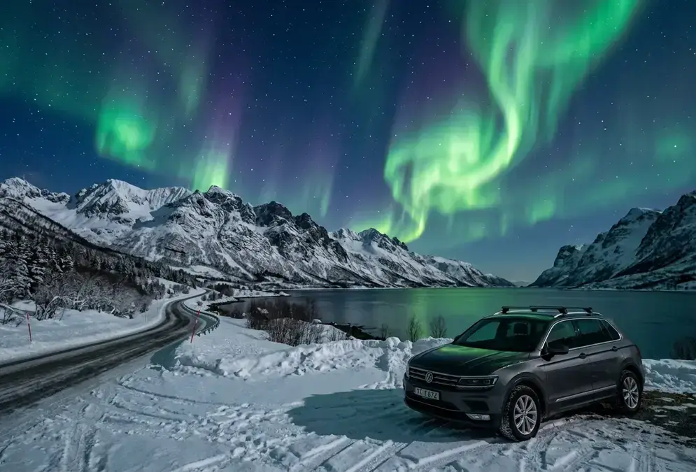 Wide-angle night shot from the E8 road south of Tromsø, Norway: green and violet aurora borealis curtains filling the sky above a snow-covered fjord valley, a lone rental car parked at a roadside viewing layby with no tour group in sight, photorealistic Arctic winter scene illustrating whether you need a tour to see northern lights in Tromsø