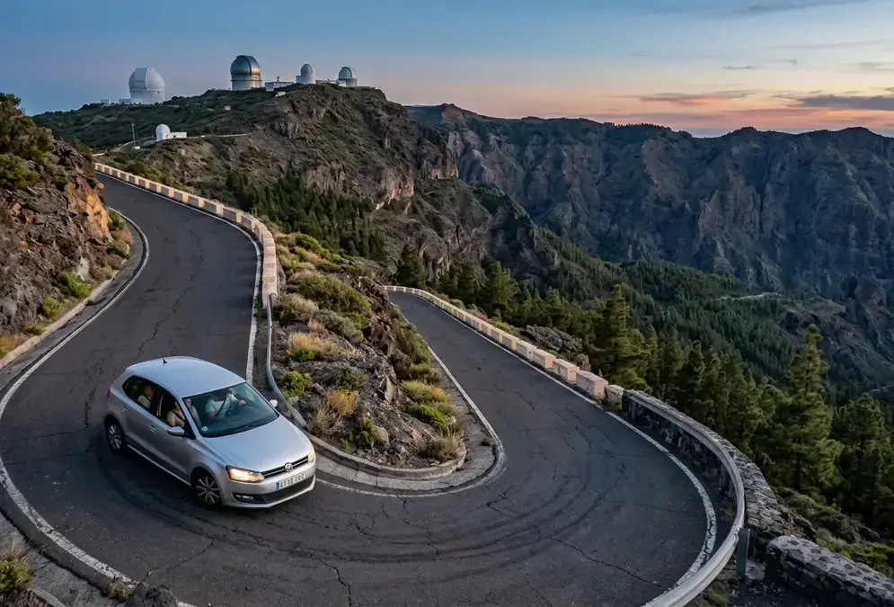 A rental car ascending the narrow, hairpin-bend summit road toward Roque de los Muchachos Observatory on La Palma at dusk, illustrating getting to La Palma's stargazing sites, with dense pine forest and a volcanic ridgeline below the observatory domes
