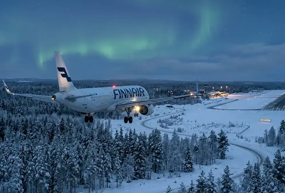 A Finnair jet on final approach into snow-covered Rovaniemi Airport at dusk, faint green aurora glow above the spruce tree line, getting to Rovaniemi Lapland by air