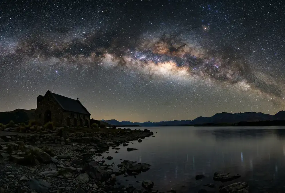 Photorealistic night view of the Church of the Good Shepherd silhouette on Lake Tekapo's shore under the Milky Way galactic core, Aoraki Mackenzie Gold Tier dark sky reserve, illustrating how many days aoraki mackenzie stargazing trips need for a clear view like this