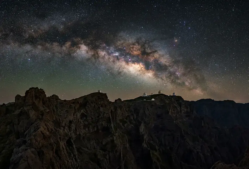 Photorealistic wide-angle nightscape of the Caldera de Taburiente rim on La Palma at full dark, Milky Way core arching above the Roque de los Muchachos Observatory domes in the distance, volcanic ridge silhouette in the foreground, no moon, Bortle 2 sky, illustrating how many days in La Palma for stargazing produce this view