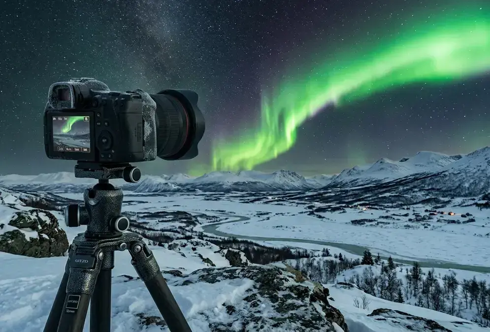 Photographer's camera on a sturdy tripod aimed at a vivid green aurora curtain above a snow-covered Arctic landscape at night, illustrating how to photograph northern lights.