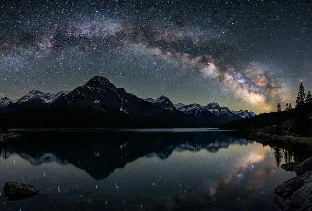 Star-filled night sky over Pyramid Lake in Jasper National Park, Alberta, with a mountain silhouette and calm water reflecting the Milky Way