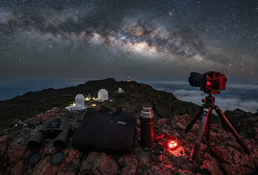 Photorealistic night scene on the Roque de los Muchachos ridge on La Palma: a red headlamp, 10x50 binoculars, a mirrorless camera on a travel tripod, a fleece jacket, and a stainless steel thermos laid out on dark volcanic rock, with the Milky Way core sharply visible overhead above the observatory domes in the distance. La Palma stargazing gear essentials for a mild but cold high-altitude night.
