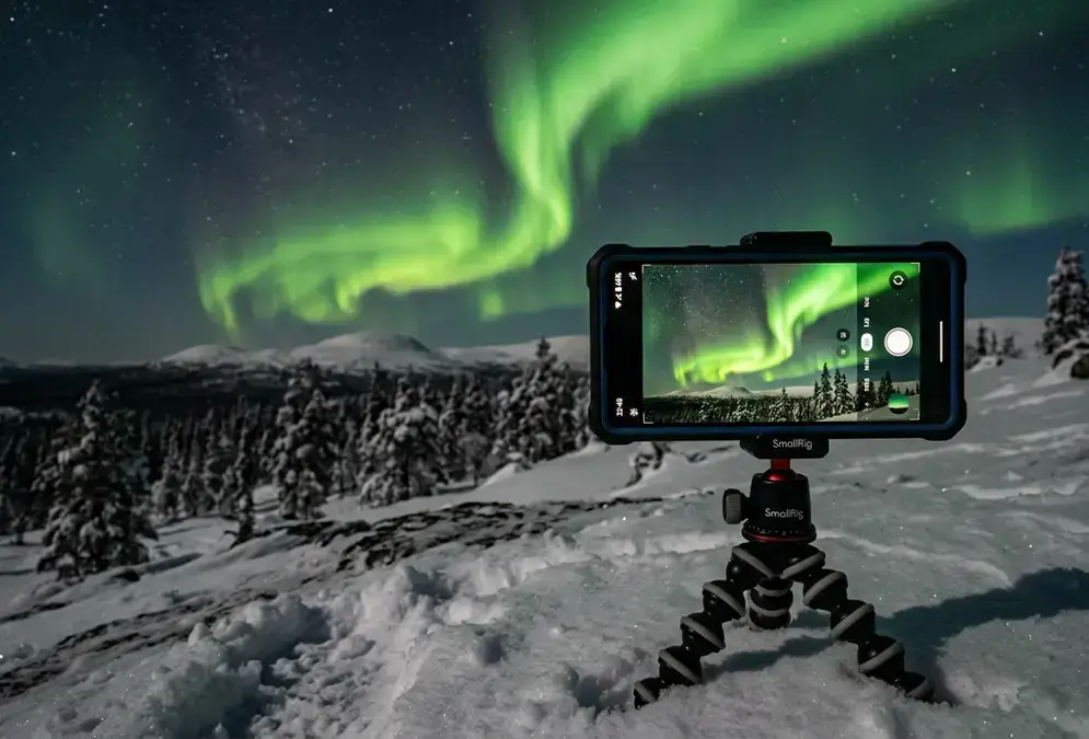 Photograph northern lights with a phone in Lapland: a smartphone mounted on a mini tripod pushed into snow, screen showing a green aurora curtain over a Finnish Lapland fell at night