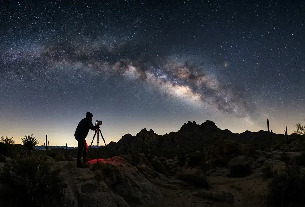 How to photograph the Milky Way: a photographer with a camera on a tripod framing the galactic core over a dark desert horizon at night, wide-angle shot, stars sharp, deep indigo sky