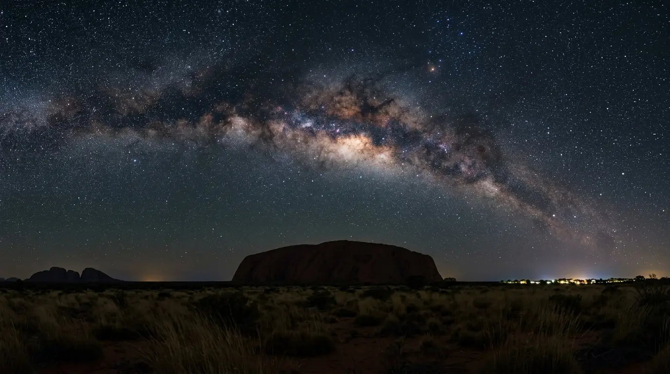 Wide dusk photograph of Uluru's silhouette beneath the Milky Way's galactic core for this Uluru stargazing FAQ, Yulara's resort lights faint on the horizon, no climbers and no close-up of sacred rock markings.